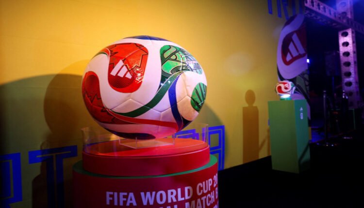 Soccer ball on a clear stand, part of a FIFA World Cup exhibit against a vivid yellow wall with other display pedestals in the background.