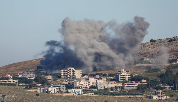 Large dark smoke plume rising behind a hillside town, with low-rise buildings in the foreground used as a backdrop for an explosion