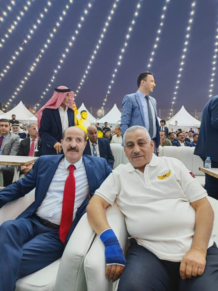 العيسوي 3 - الصافي نيوز Two men in formal attire sit on a white sofa at an outdoor evening event under string lights, with the crowd and white tents in the background.
