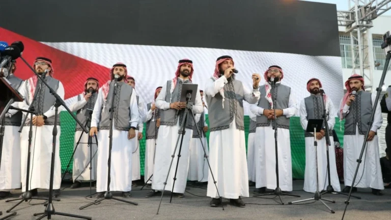 العيسوي 5 - الصافي نيوز Group of men in white thobes and gray vests singing on a stage with multiple microphones and stands, wearing red-and-white keffiyehs.