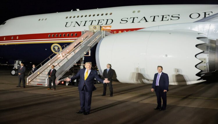 Man in a dark suit with a yellow tie stands on a tarmac, speaking beside a large presidential aircraft with boarding stairs at night.