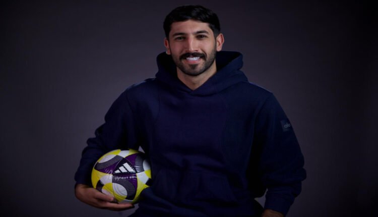 Portrait of a man in a navy hoodie holding a yellow and purple soccer ball against a dark backdrop, smiling at the camera.